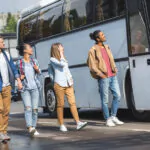 cheerful multiethnic tourists with backpacks walking near travel bus at street