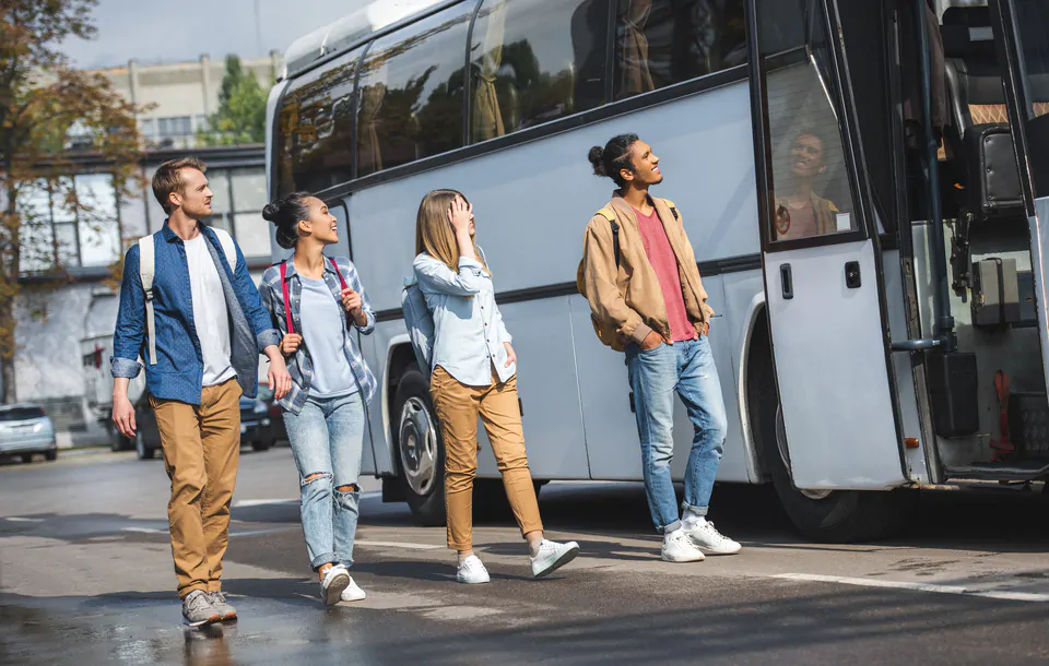 cheerful multiethnic tourists with backpacks walking near travel bus at street